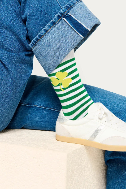 Person wearing green and white striped socks with a yellow clover design, paired with blue jeans and white sneakers.