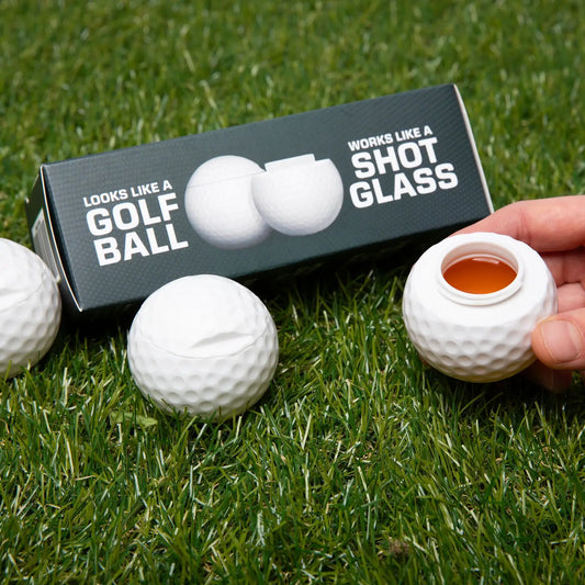 Golf ball-shaped shot glass on grass with packaging and additional golf balls.