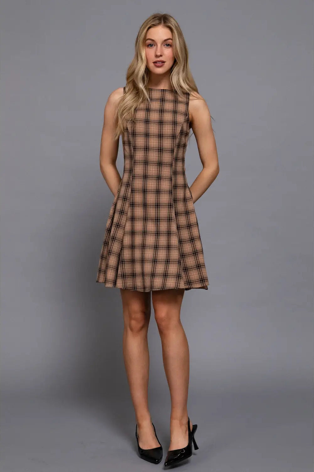 A woman modeling a sleeveless, knee-length plaid dress in brown and beige tones, paired with black heels, against a solid gray studio background.
