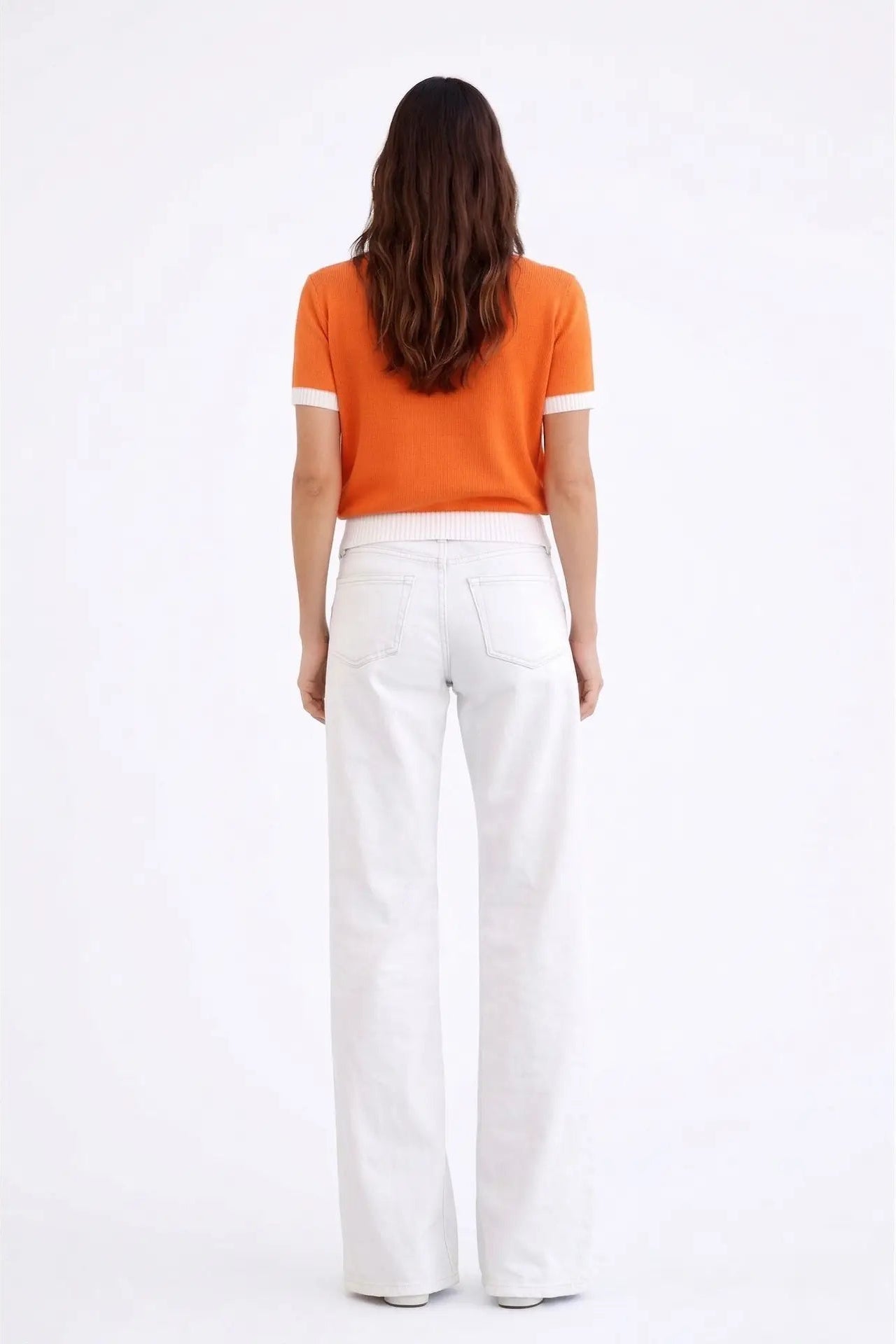 A woman viewed from behind wearing a bright orange short-sleeve knit top with white sleeve trim and white wide-leg trousers, standing against a plain white studio background.