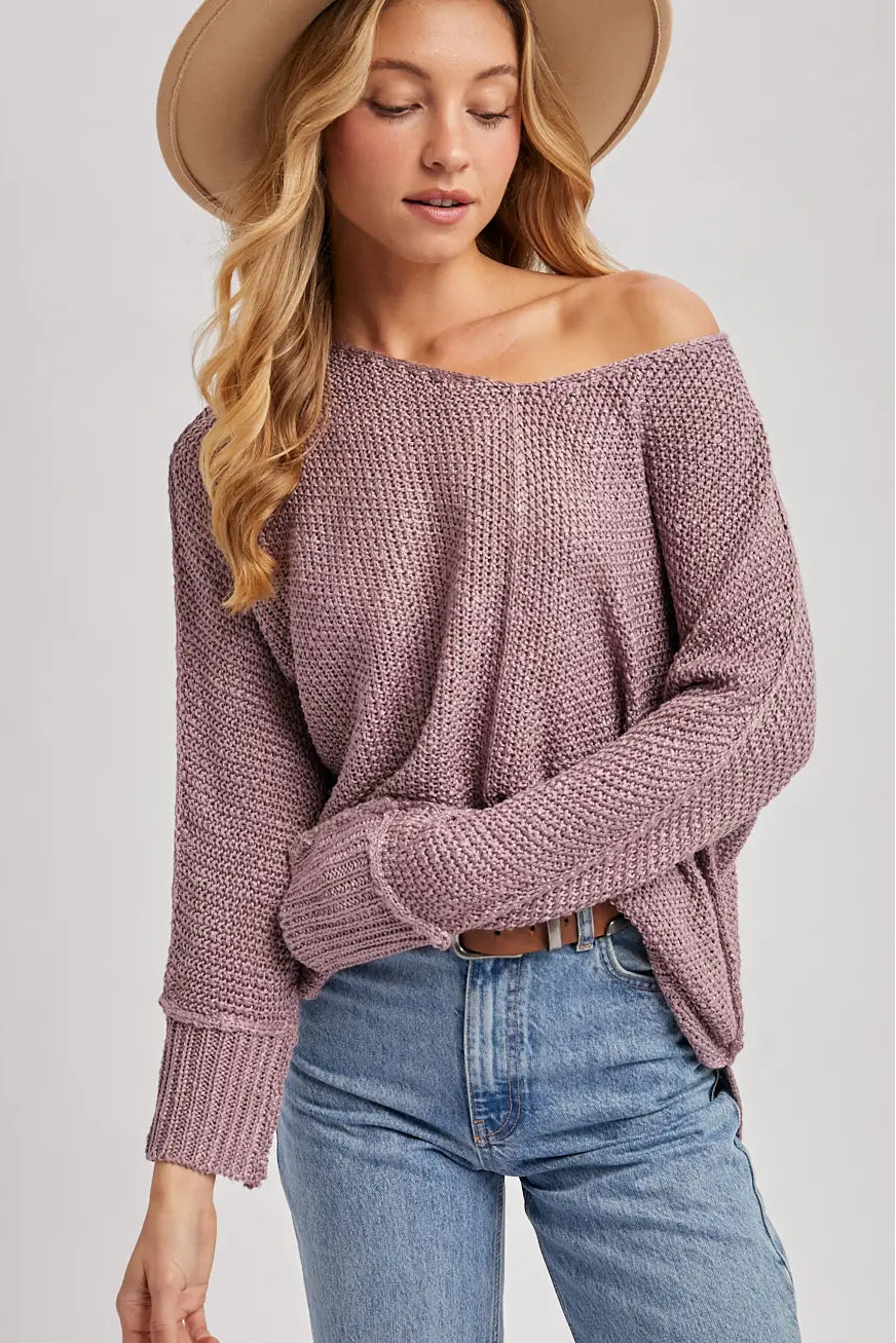 A woman wearing a chunky knit off-the-shoulder sweater in dusty mauve, paired with blue jeans and a wide-brim tan hat, against a light gray studio background.