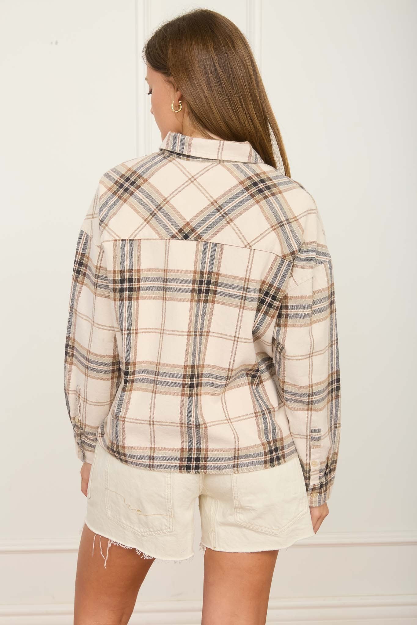 A woman shown from the back wearing a cream-colored plaid button-up shirt with brown and black lines, paired with off-white shorts, against a white paneled wall.