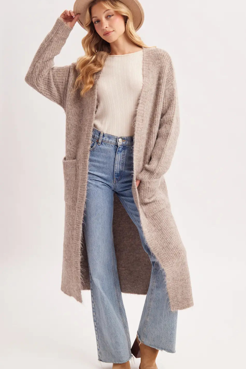 A woman modeling a long, open-front, chunky knit cardigan in a light taupe-beige color, styled with a cream ribbed top, light-wash blue jeans, and a tan hat against a plain white studio background.