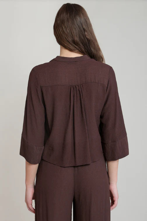 A woman viewed from the back wearing a dark brown cropped top with three-quarter sleeves and matching wide-leg trousers against a light gray studio background.