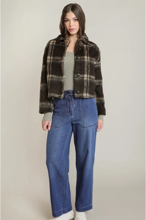 A model stands against a light beige studio background wearing a brown and black plaid cropped jacket over a sage green top, paired with blue wide-leg jeans and white sneakers.