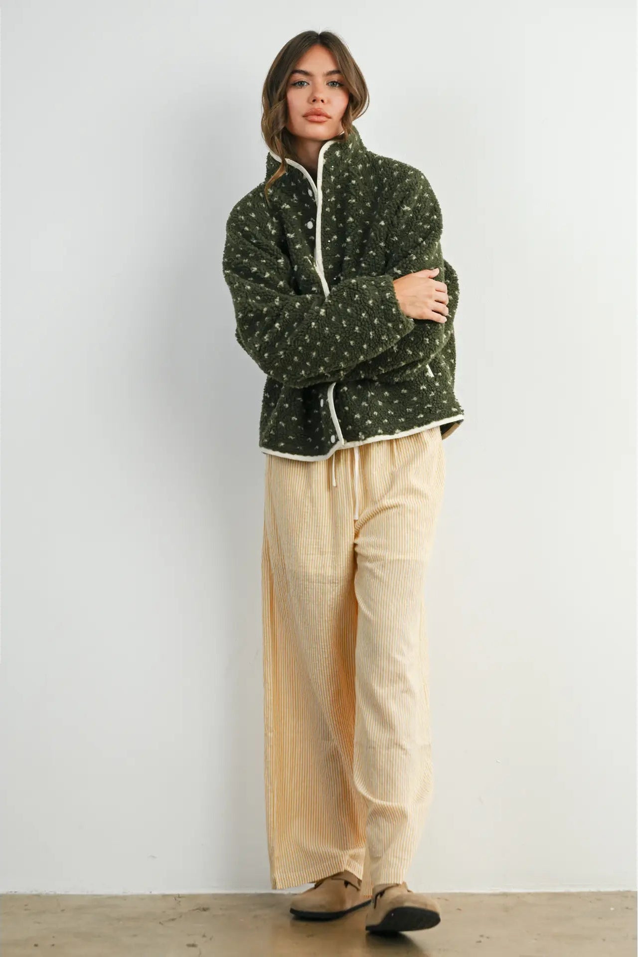 A woman stands against a plain light gray wall wearing a dark green quilted jacket with a small white floral pattern and white trim, paired with loose beige pants and tan shoes.