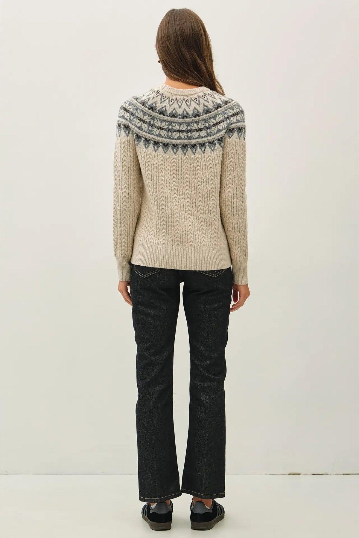 A woman viewed from behind wearing a cream-colored cable-knit sweater with a gray and blue geometric Fair Isle yoke pattern, paired with dark wash jeans and black shoes, against a plain white studio background.