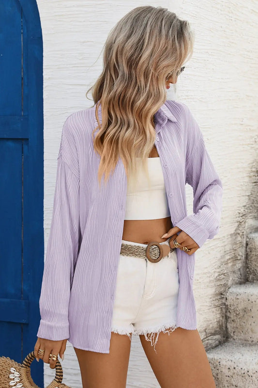 Woman wearing a light purple shirt over a white crop top and shorts, standing against a white wall with a blue door.