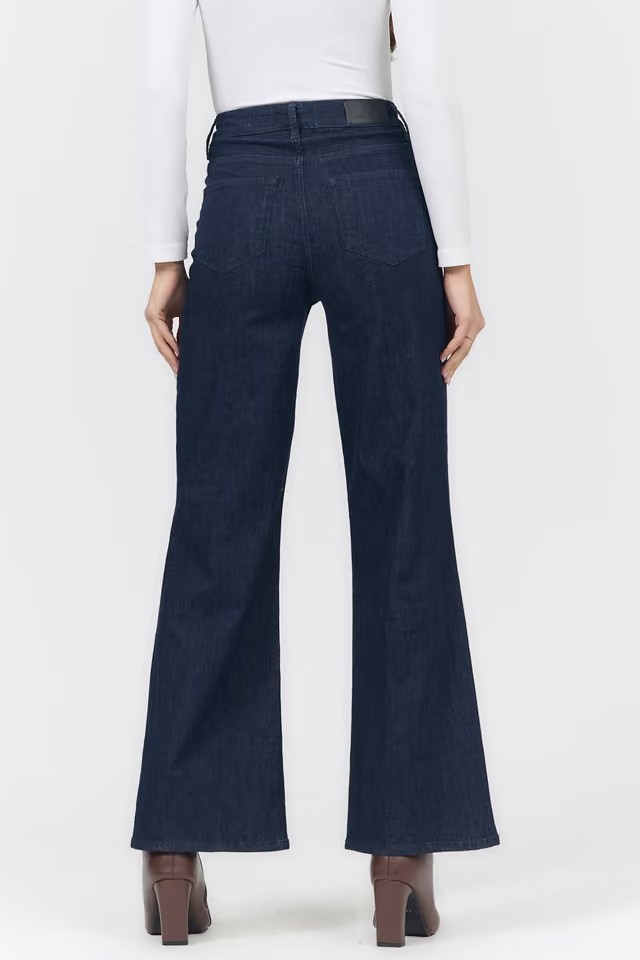 Rear view of a person wearing dark blue wide-leg flare jeans with a white long-sleeve top and brown heeled shoes against a light gray studio background.