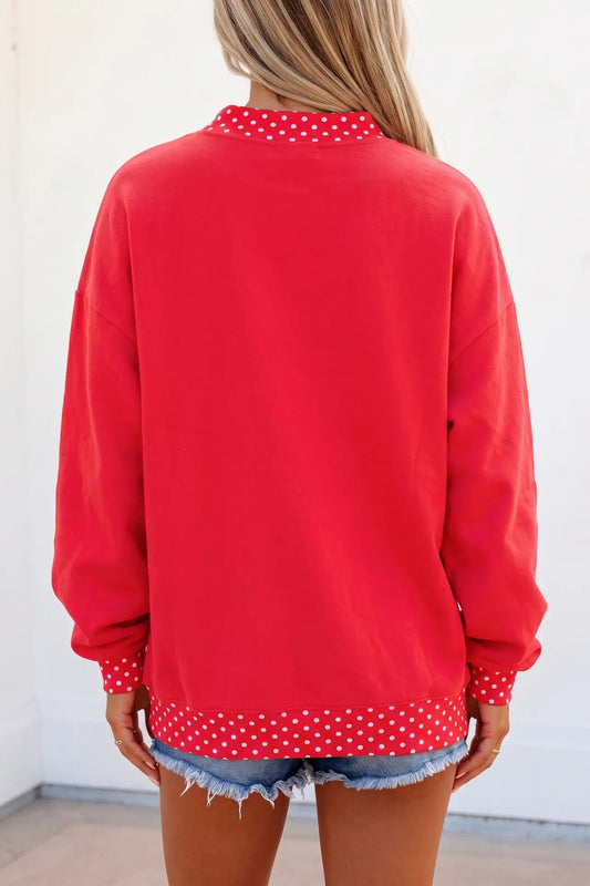 A woman shown from the back wearing a bright red long-sleeve sweatshirt with white polka dot trim at the collar, cuffs, and hem, paired with denim shorts.