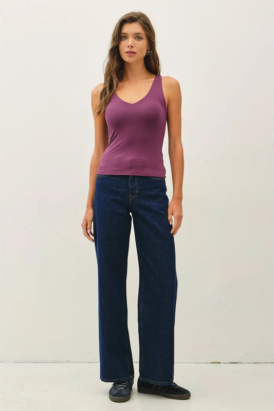 A woman modeling a sleeveless purple V-neck tank top and dark blue wide-leg jeans against a plain off-white studio background.