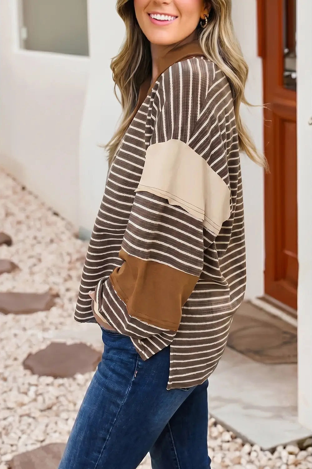 A woman wearing a brown and beige striped long-sleeve top with solid beige and brown color-block panels, paired with blue jeans, standing in front of a white wall and wooden door.