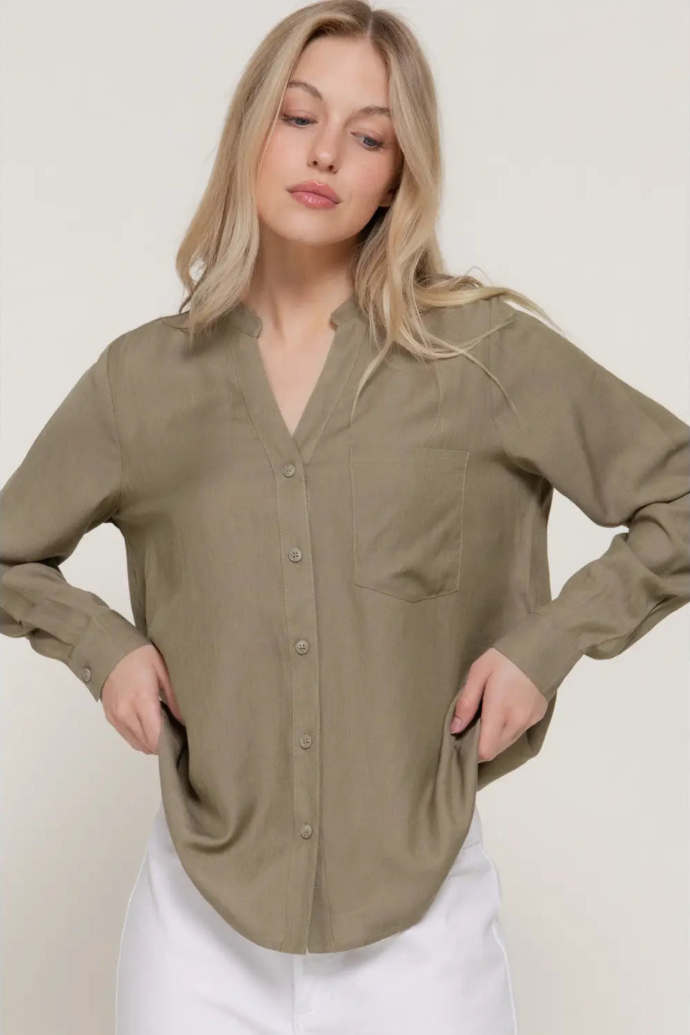 A woman wearing a relaxed-fit, long-sleeve button-up shirt in a muted olive khaki color, paired with white pants, set against a plain light beige studio background.
