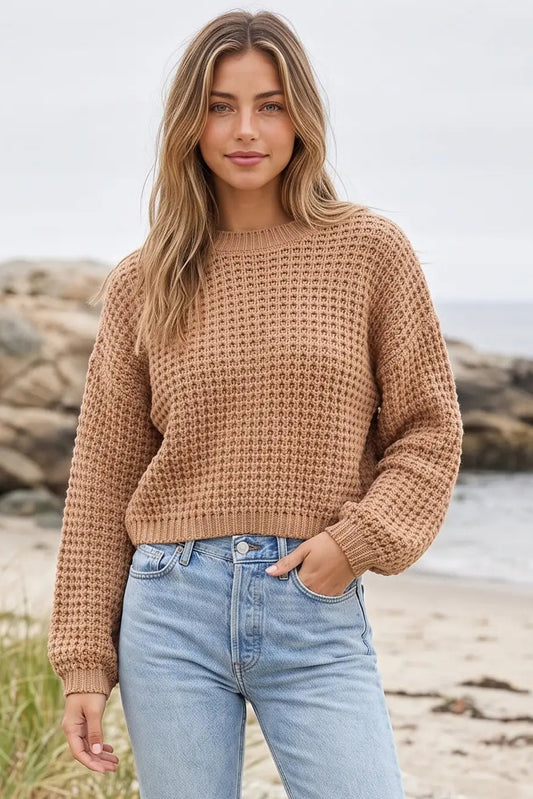 A woman standing on a beach wearing a textured, cropped waffle-knit sweater in a warm camel brown color, paired with light-wash blue jeans.