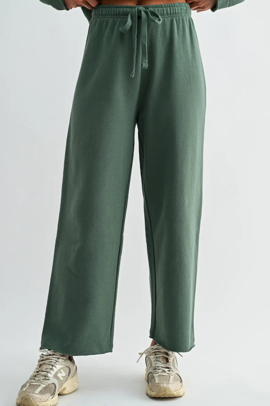 A person wearing wide-leg sage green sweatpants with an elastic drawstring waist, paired with beige sneakers, shown from the waist down against a light gray studio background.