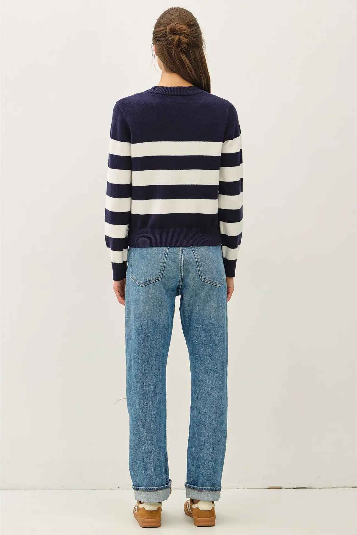 A woman viewed from behind wearing a navy and white horizontally striped long-sleeve knit sweater, light-wash blue jeans, and tan wedge sandals against a plain off-white studio background.