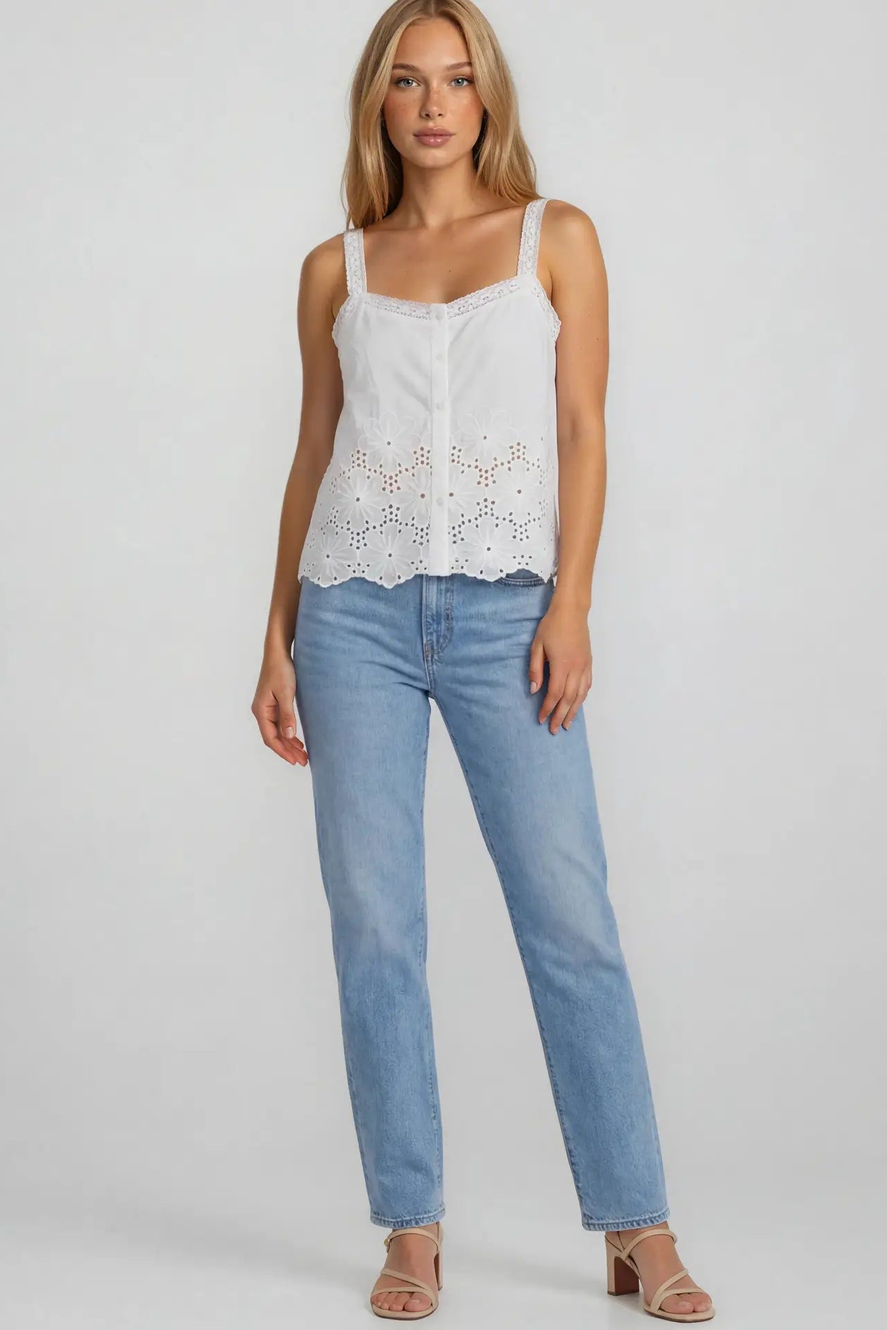 Woman wearing a white sleeveless top with lace details and light blue jeans on a plain background