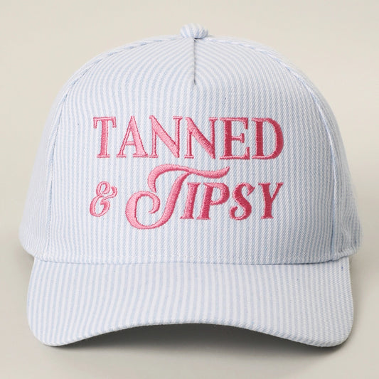 Light blue cap with pink 'Tanned & Tipsy' text on a beige background