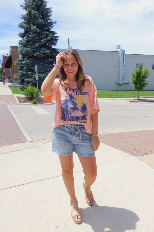 Woman in a pink graphic t-shirt and denim shorts standing on a sidewalk with trees and buildings in the background.