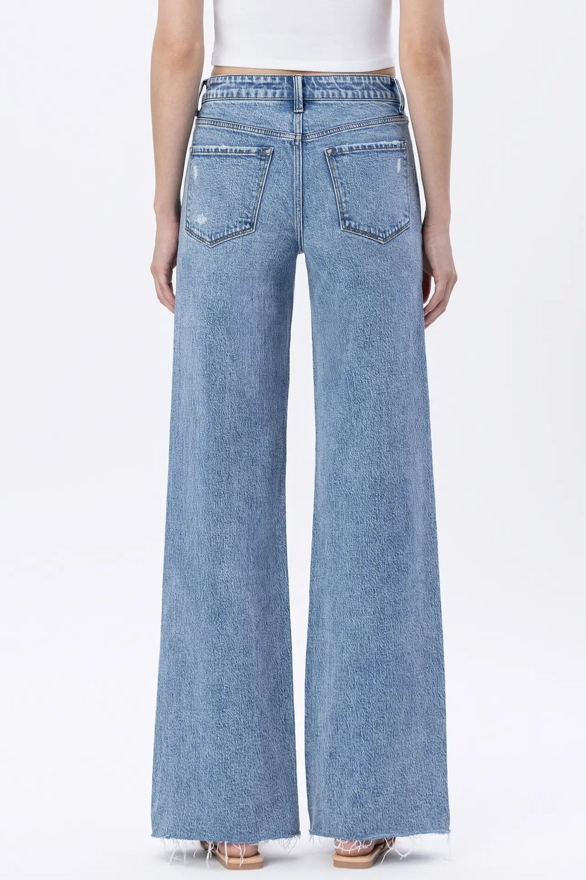 A model shown from the back wearing light blue wide-leg denim jeans with a frayed hem, paired with a white top and beige sandals, against a plain white studio background.