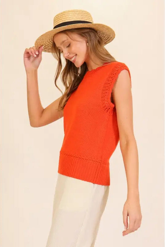 A woman wearing a bright orange sleeveless knit top with textured lace-up side details, paired with cream pants and a straw boater hat, against a solid beige background.