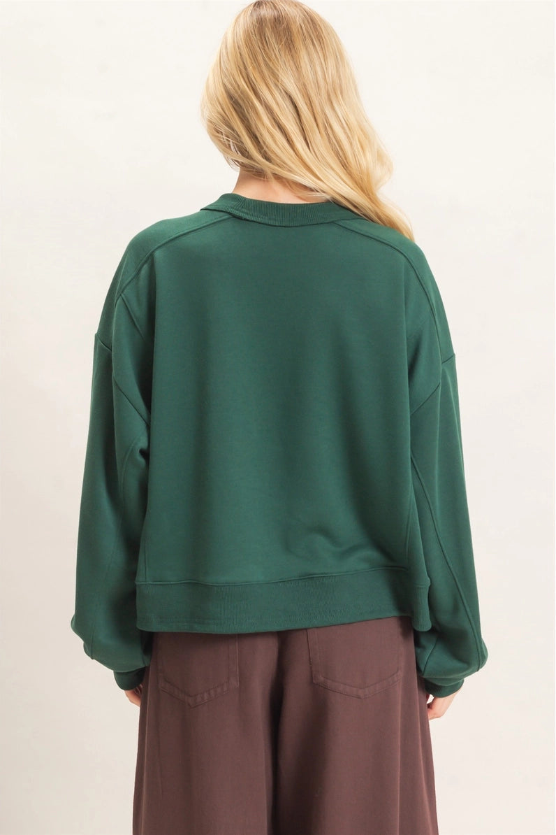 A woman viewed from behind wearing a dark green cropped sweatshirt and brown wide-leg pants against a plain off-white background.