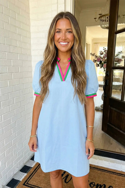 Woman wearing a light blue dress with pink and green trim in a home setting.