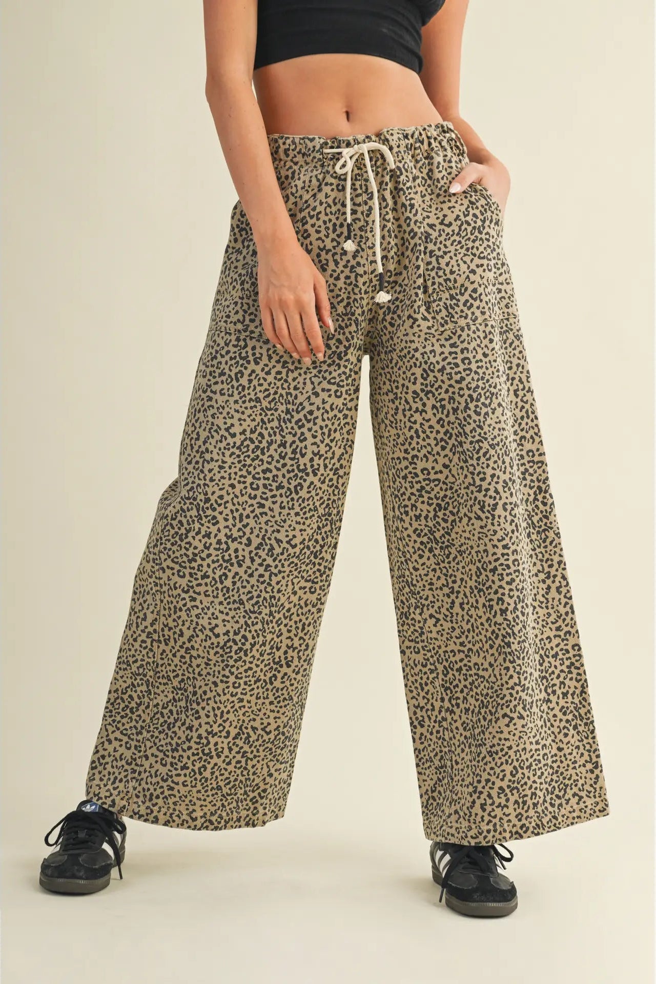 A person wearing loose, wide-leg leopard-print pants with a cream drawstring waist, paired with a black crop top and black sneakers, against a plain beige studio background.