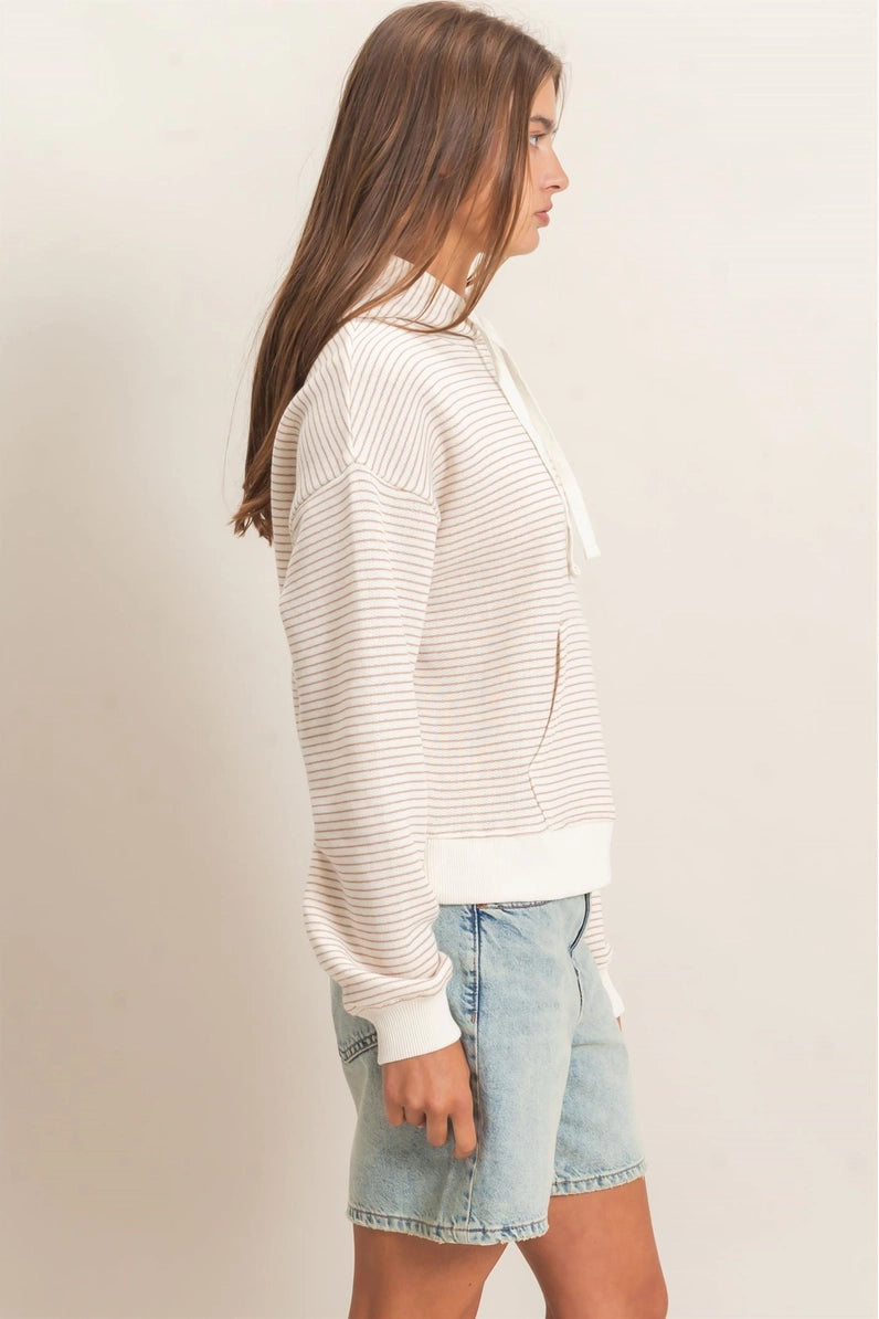 A woman shown in side profile wearing a cream and white horizontally striped knit hoodie with a drawstring, paired with light-wash denim shorts, against a plain beige studio background.