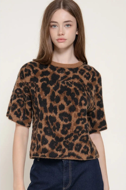 A woman wearing a brown and black leopard-print short-sleeve knit top with dark blue jeans, photographed against a plain light beige studio background.