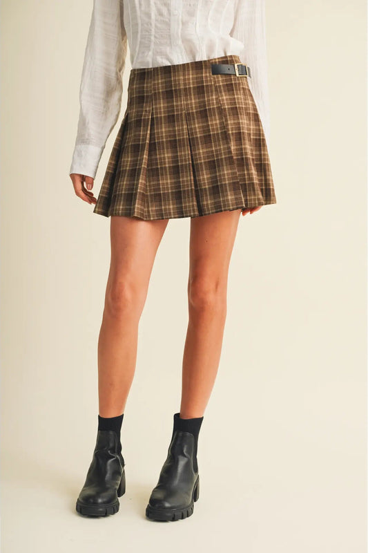 A model wearing a brown and beige plaid pleated mini skirt with a black belt, paired with a white shirt and black ankle boots, against a solid cream background.