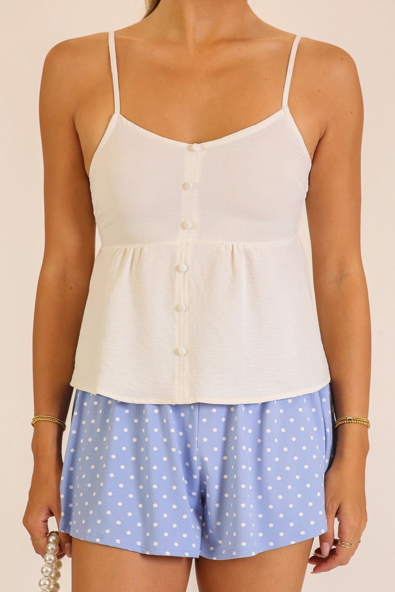 A woman wearing a white camisole top with thin straps and a button front, paired with light blue shorts with white polka dots, against a plain beige background.