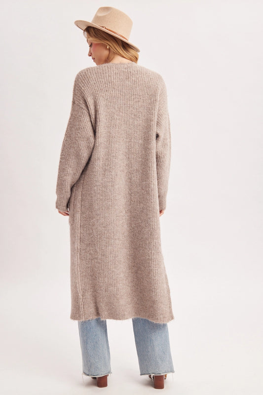A woman viewed from behind wearing a long, ribbed knit cardigan in a light taupe-beige color, styled with light-wash blue jeans and a beige wide-brim hat against a plain white studio background.