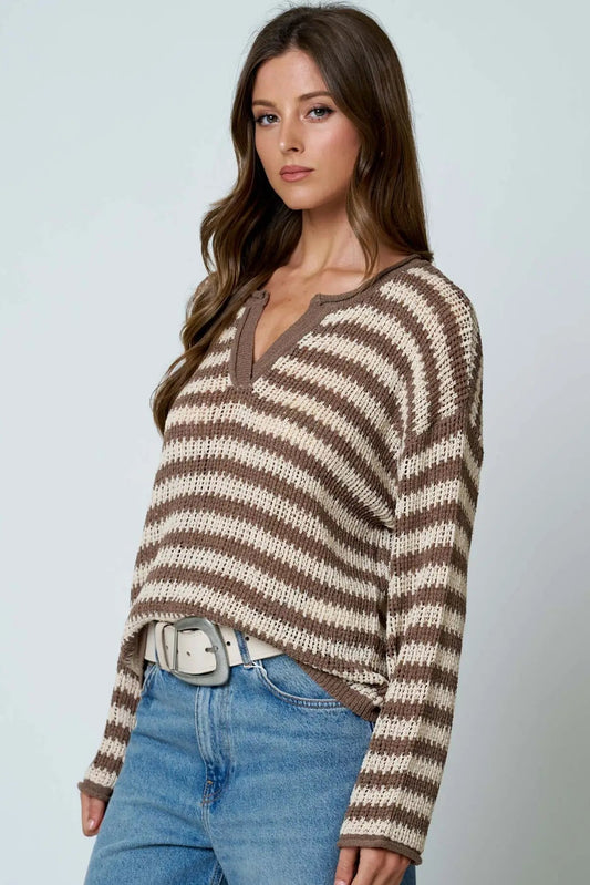 A woman wearing a brown and cream striped knit sweater with a split V-neckline, paired with blue jeans and a silver belt, against a light gray studio background.