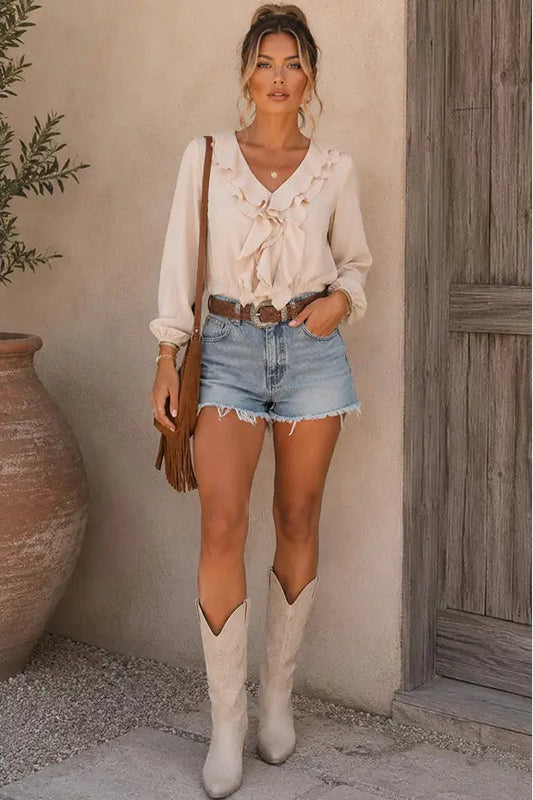 A woman stands outdoors wearing a cream ruffled long-sleeve blouse, light blue denim shorts, a brown belt, and off-white cowboy boots, with a brown shoulder bag.