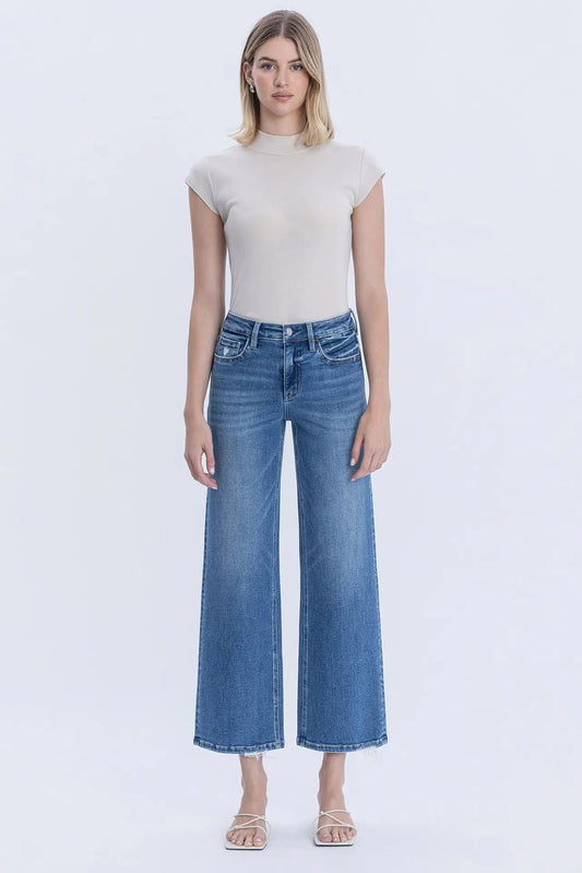 A woman modeling a beige short-sleeve mock neck top and blue wide-leg cropped jeans against a plain white studio background.