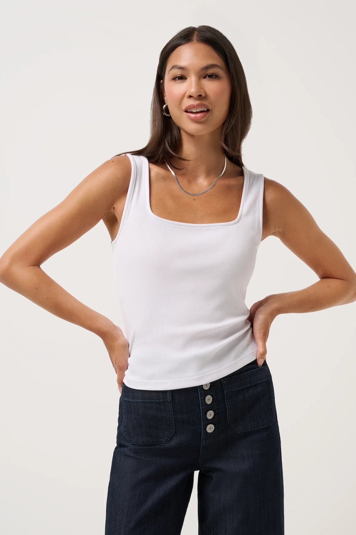 Woman wearing a white tank top and dark blue jeans against a plain background