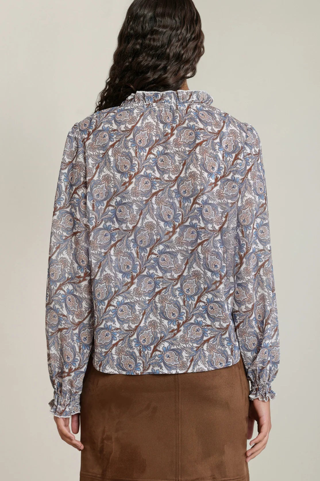 A woman shown from the back wearing a long-sleeve button-up blouse with an intricate paisley pattern in blue, beige, and brown, paired with a brown skirt, against a neutral studio background.