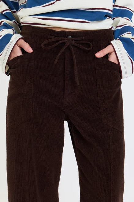 A close-up of a person wearing dark brown corduroy pants with a drawstring waist and side pockets, paired with a blue, white, and burgundy striped long-sleeve top.