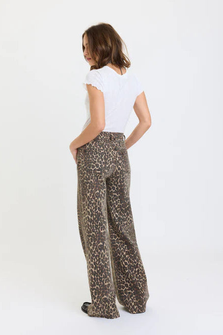 A woman shown from the back wearing wide-leg leopard-print trousers and a white short-sleeve top, standing against a plain white studio background.