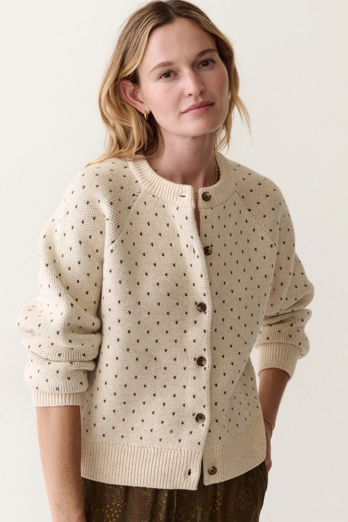 A woman wearing a cream-colored knit cardigan with small dark brown polka dots and dark brown buttons, photographed against a plain off-white studio background.