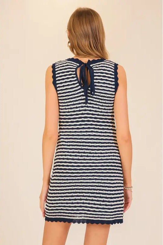 A woman shown from the back wearing a sleeveless knit dress with horizontal navy and off-white stripes, a back tie detail, and scalloped edges, set against a plain beige background.