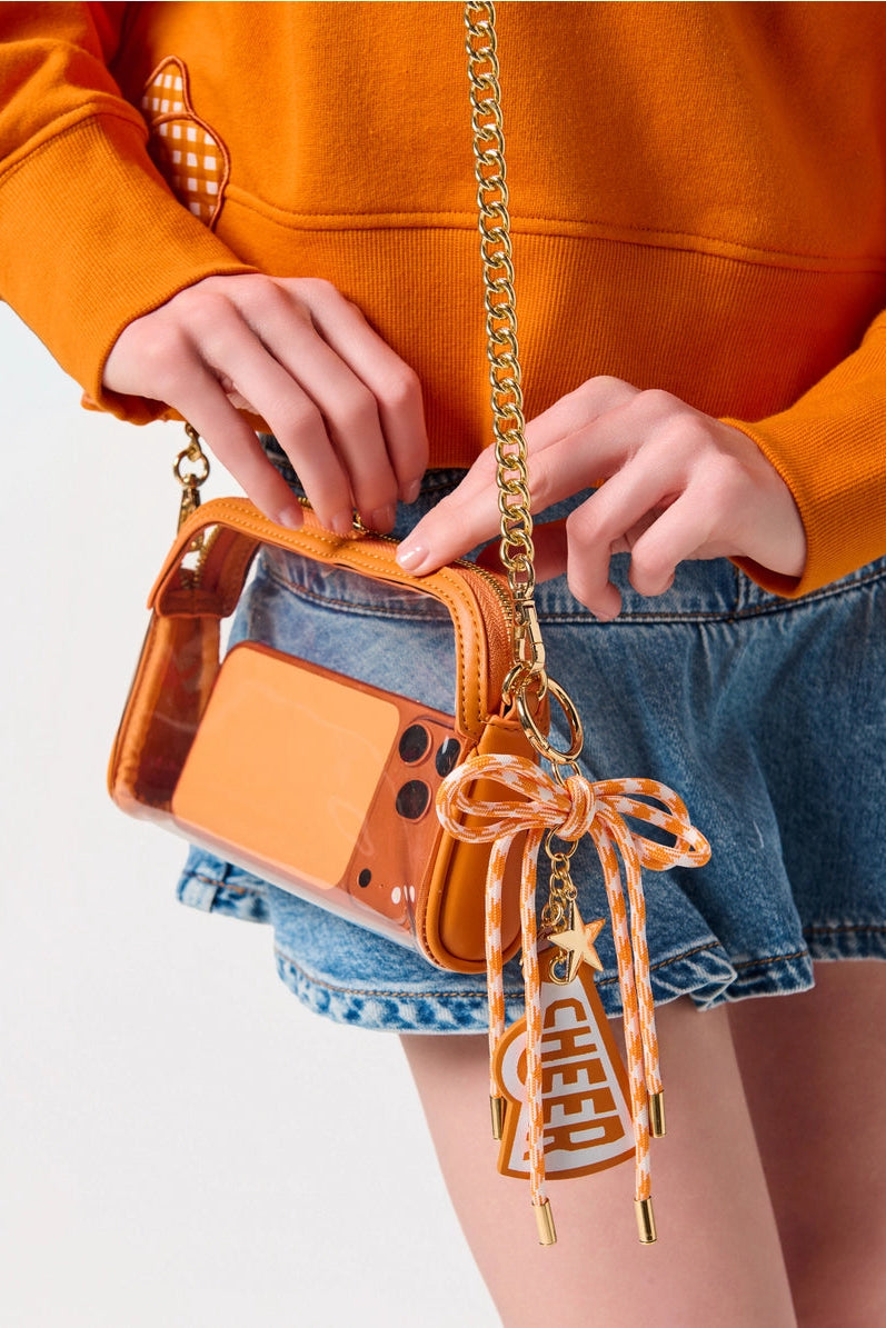 A close-up of a person wearing an orange hoodie and denim skirt, carrying a small transparent orange bag with a gold chain strap and a decorative orange-and-white chevron ribbon charm.