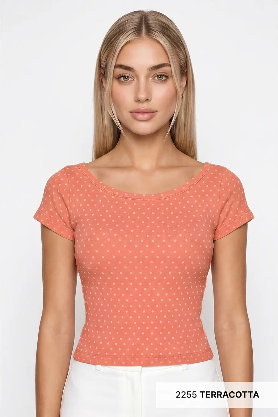A woman modeling a terracotta short-sleeve scoop-neck top with a small white geometric pattern, paired with white pants against a light gray background.