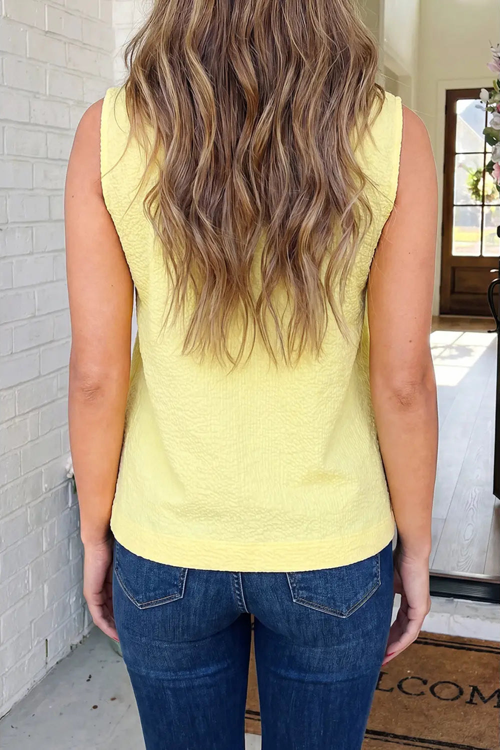 Person wearing a yellow sleeveless top and blue jeans, standing in front of a door.