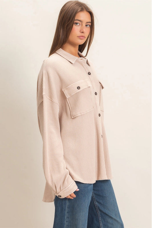 A woman wearing a light beige ribbed long-sleeve button-up shirt with chest pockets and blue jeans, standing against a plain light beige background.