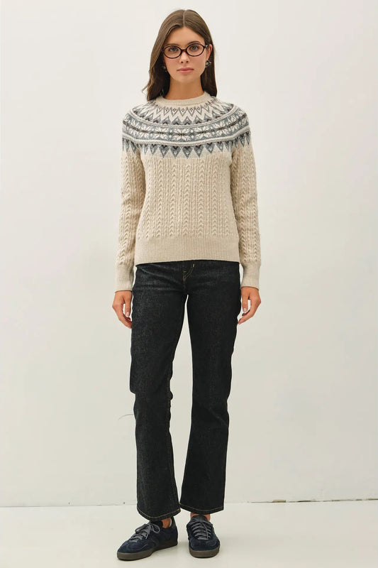A woman wearing a cream cable-knit sweater with a gray and white Fair Isle yoke pattern, paired with dark wash jeans and navy sneakers, standing against a plain off-white studio background.