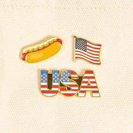 Three pins featuring a hot dog, American flag, and 'USA' text on a textured beige background.