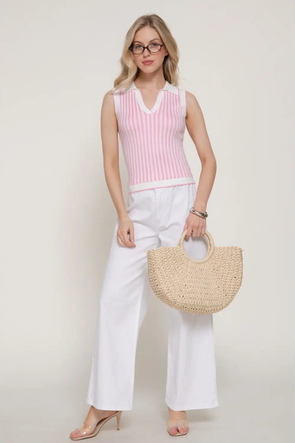 Woman wearing a pink sleeveless top and white pants, holding a straw bag on a plain background