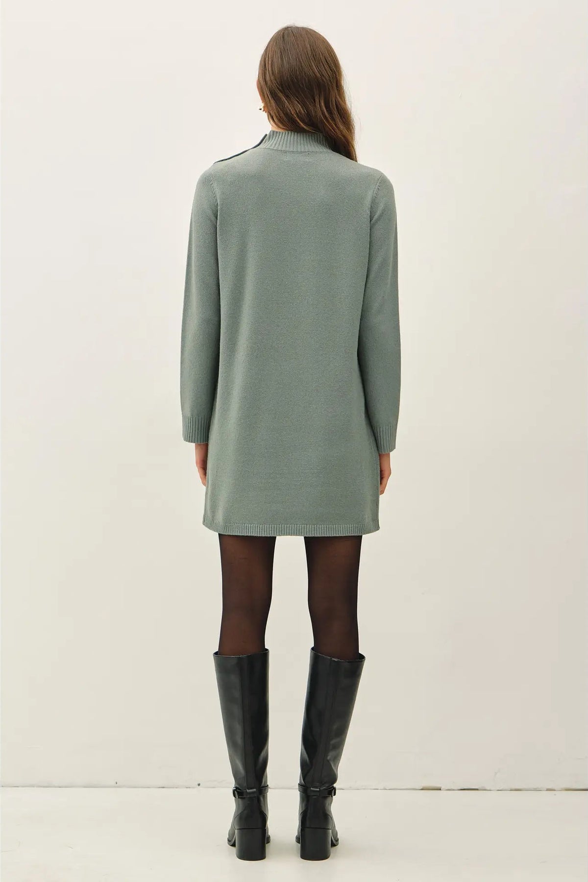 A woman viewed from the back wearing a sage green long-sleeve knit dress with black tights and black knee-high boots, standing against a plain off-white studio background.